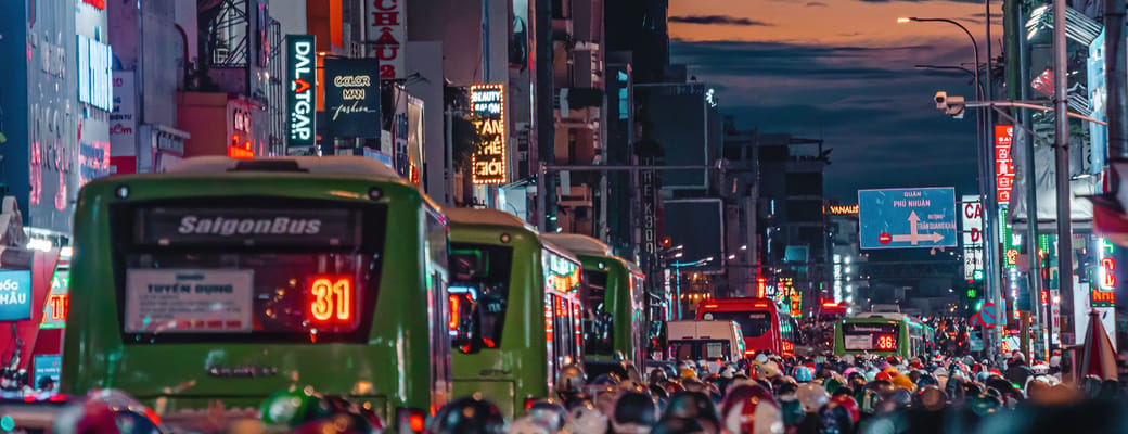 Saigon Vespa-tur gennem travle vietnamesiske gader om aftenen med neonskilt og gr&oslash;n SaigonBus i baggrunden