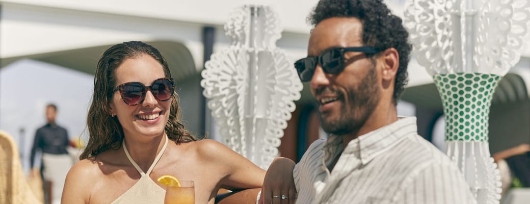 Par på Celebrity Cruises dæk nyder drinks og snacks sammen, mand og kvinde smiler i solskin med skibets faciliteter i baggrunden