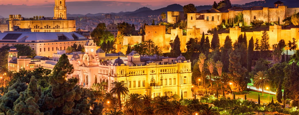 Aftenstemning over Málaga med oplyste historiske bygninger, palmer og rosa himmel
