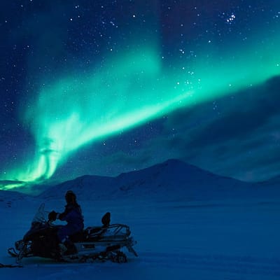 Snescooter p&aring; tur i den bl&aring; time med gr&oslash;nt norslys p&aring; himlen