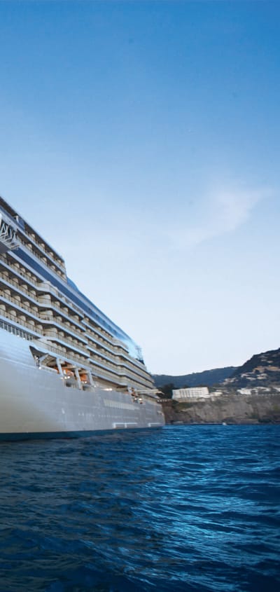Krydstogtskibet Seven Seas Explorer set fra stævnen med kyst og klipper i baggrunden