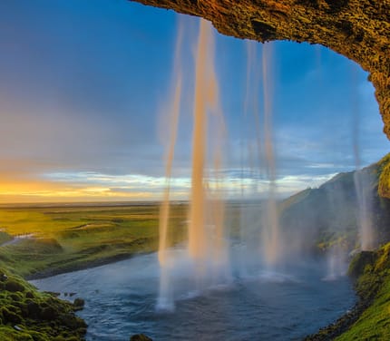 Seljalandsfoss vandfald p&aring; Island ved solnedgang med gyldent lys gennem vandfaldet og gr&oslash;nne marker i baggrunden