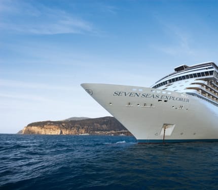 Krydstogtskibet Seven Seas Explorer set fra st&aelig;vnen med kyst og klipper i baggrunden