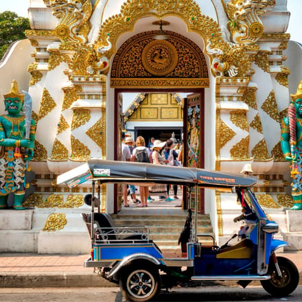 Traditionel bl&aring; tuk-tuk parkeret foran farverigt thailandsk tempel med gulddetaljer og gr&oslash;nne tempelvogter-statuer