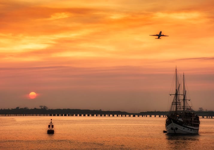 Solnedgang over Benoa havn på Bali med sejlskib i forgrunden og fly, der letter i horisonten