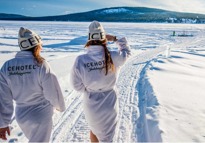 to kvinder i hvide badekåber og huer går i sneen ved Icehotel i svensk lapland